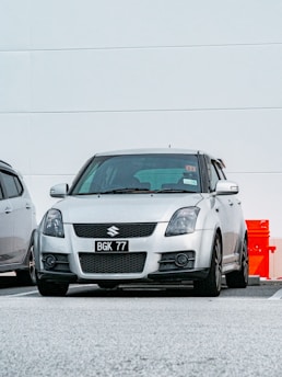 A silver Suzuki car is parked in a parking lot. The vehicle has a license plate number BGK 77 and is located next to another parked car. The background features a white wall with minimal detailing, and there is a red object partially visible to the right.
