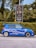 A blue car with sporty decals is parked on the street next to a green hedge. Behind the car, a black signboard advertises real estate, surrounded by lush green foliage and trees.