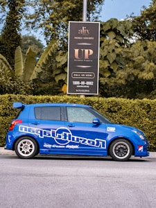 A blue car with sporty decals is parked on the street next to a green hedge. Behind the car, a black signboard advertises real estate, surrounded by lush green foliage and trees.