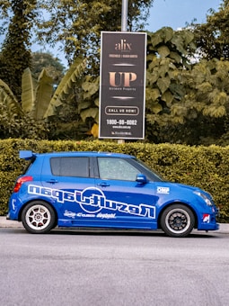 A blue car with sporty decals is parked on the street next to a green hedge. Behind the car, a black signboard advertises real estate, surrounded by lush green foliage and trees.