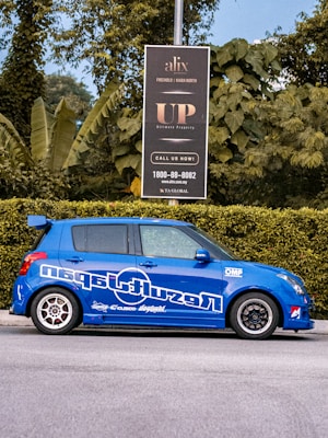 A blue car with sporty decals is parked on the street next to a green hedge. Behind the car, a black signboard advertises real estate, surrounded by lush green foliage and trees.