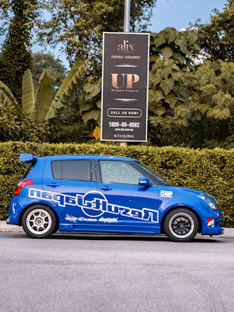 A blue car with sporty decals is parked on the street next to a green hedge. Behind the car, a black signboard advertises real estate, surrounded by lush green foliage and trees.