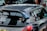 A heavily customized car with numerous stickers covering the rear window, signifying various automotive brands and modifications. The car features a prominent rear spoiler and a dark exterior. The setting includes other parked cars in an urban environment.