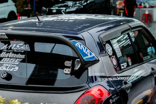 A heavily customized car with numerous stickers covering the rear window, signifying various automotive brands and modifications. The car features a prominent rear spoiler and a dark exterior. The setting includes other parked cars in an urban environment.