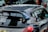 A heavily customized car with numerous stickers covering the rear window, signifying various automotive brands and modifications. The car features a prominent rear spoiler and a dark exterior. The setting includes other parked cars in an urban environment.