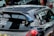 A heavily customized car with numerous stickers covering the rear window, signifying various automotive brands and modifications. The car features a prominent rear spoiler and a dark exterior. The setting includes other parked cars in an urban environment.