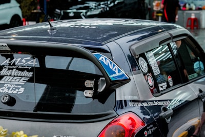 A heavily customized car with numerous stickers covering the rear window, signifying various automotive brands and modifications. The car features a prominent rear spoiler and a dark exterior. The setting includes other parked cars in an urban environment.