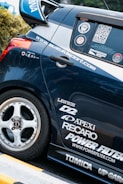 A close-up view of the rear section of a sporty car adorned with multiple brand decals including Apex, Recaro, and Power Filter. The car is navy blue with a silver stripe and features custom wheels.
