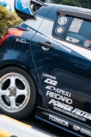 A close-up view of the rear section of a sporty car adorned with multiple brand decals including Apex, Recaro, and Power Filter. The car is navy blue with a silver stripe and features custom wheels.