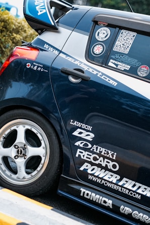 A close-up view of the rear section of a sporty car adorned with multiple brand decals including Apex, Recaro, and Power Filter. The car is navy blue with a silver stripe and features custom wheels.