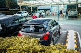 A compact car with various stickers on the back is parked at a car wash location. Several people are seen around, with one person apparently preparing their vehicle for service. The background includes multiple cars being washed under a well-lit canopy.