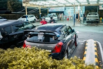 A compact car with various stickers on the back is parked at a car wash location. Several people are seen around, with one person apparently preparing their vehicle for service. The background includes multiple cars being washed under a well-lit canopy.