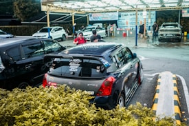 A compact car with various stickers on the back is parked at a car wash location. Several people are seen around, with one person apparently preparing their vehicle for service. The background includes multiple cars being washed under a well-lit canopy.