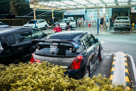 A compact car with various stickers on the back is parked at a car wash location. Several people are seen around, with one person apparently preparing their vehicle for service. The background includes multiple cars being washed under a well-lit canopy.