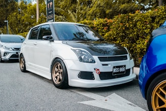 A modified white car with a black hood is parked on the side of a street. It features bronze alloy wheels and a red tow hook on the front bumper. The vehicle is aligned next to another car, with greenery and trees in the background.
