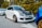 A modified white car with a black hood is parked on the side of a street. It features bronze alloy wheels and a red tow hook on the front bumper. The vehicle is aligned next to another car, with greenery and trees in the background.