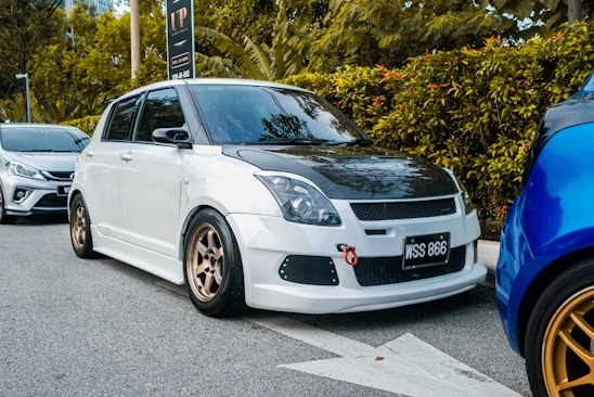 A modified white car with a black hood is parked on the side of a street. It features bronze alloy wheels and a red tow hook on the front bumper. The vehicle is aligned next to another car, with greenery and trees in the background.