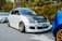 A modified white car with a black hood is parked on the side of a street. It features bronze alloy wheels and a red tow hook on the front bumper. The vehicle is aligned next to another car, with greenery and trees in the background.