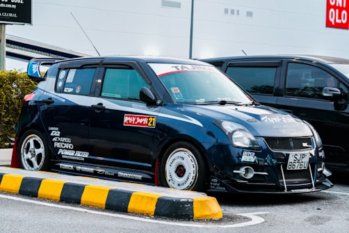 A modified blue car is parked in a lot, featuring various decals and stickers from brands such as Recaro, Power Filter, and others. The vehicle sports custom rims, a body kit, and a rear spoiler. The environment suggests a commercial area with a Uniqlo store visible in the background.