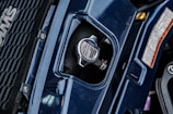 A close-up view of a vehicle's radiator cap, surrounded by dark blue bodywork and a portion of a grille with a logo on the left. There are detailed safety labels on the right, and the metal cap features instructions and branding.