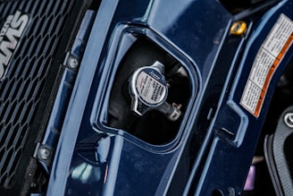 A close-up view of a vehicle's radiator cap, surrounded by dark blue bodywork and a portion of a grille with a logo on the left. There are detailed safety labels on the right, and the metal cap features instructions and branding.
