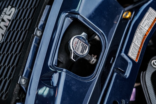 A close-up view of a vehicle's radiator cap, surrounded by dark blue bodywork and a portion of a grille with a logo on the left. There are detailed safety labels on the right, and the metal cap features instructions and branding.