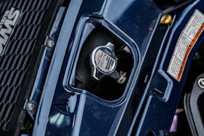 A close-up view of a vehicle's radiator cap, surrounded by dark blue bodywork and a portion of a grille with a logo on the left. There are detailed safety labels on the right, and the metal cap features instructions and branding.