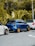 A row of three compact cars is parked on a street, with a lush green forest in the background. The central car is blue with gold wheels, while the two adjacent cars are white. The scene features a tropical environment with large leafy plants.