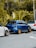 A row of three compact cars is parked on a street, with a lush green forest in the background. The central car is blue with gold wheels, while the two adjacent cars are white. The scene features a tropical environment with large leafy plants.