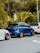 A row of three compact cars is parked on a street, with a lush green forest in the background. The central car is blue with gold wheels, while the two adjacent cars are white. The scene features a tropical environment with large leafy plants.