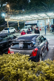 A car wash facility with several cars and a few people present. A Suzuki Swift with various stickers and decals is prominently parked in the foreground. The background features a sheltered area with signs displaying services offered at the car wash. The setting is partially obscured by bushes, and the lighting suggests it is either late afternoon or evening.