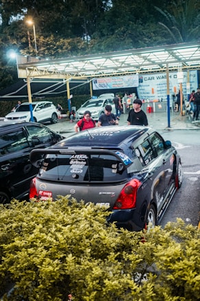 A car wash facility with several cars and a few people present. A Suzuki Swift with various stickers and decals is prominently parked in the foreground. The background features a sheltered area with signs displaying services offered at the car wash. The setting is partially obscured by bushes, and the lighting suggests it is either late afternoon or evening.