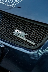 A close-up view of a car's front grille featuring the logo SWK Suzuki Works Kurume. The grille has a honeycomb pattern, and the surrounding areas of the car are a glossy blue color with visible reflections.