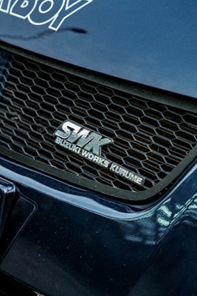 A close-up view of a car's front grille featuring the logo SWK Suzuki Works Kurume. The grille has a honeycomb pattern, and the surrounding areas of the car are a glossy blue color with visible reflections.