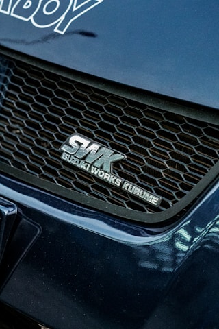 A close-up view of a car's front grille featuring the logo SWK Suzuki Works Kurume. The grille has a honeycomb pattern, and the surrounding areas of the car are a glossy blue color with visible reflections.