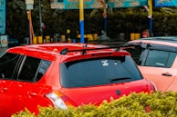 A parking lot or car wash area with two cars parked next to each other, one red and the other a lighter color. The red car has tinted windows, a roof rack, and a sticker on the back window. There are colorful signs and poles in the background with bushes in the foreground.