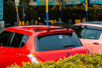 A parking lot or car wash area with two cars parked next to each other, one red and the other a lighter color. The red car has tinted windows, a roof rack, and a sticker on the back window. There are colorful signs and poles in the background with bushes in the foreground.