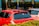 A parking lot or car wash area with two cars parked next to each other, one red and the other a lighter color. The red car has tinted windows, a roof rack, and a sticker on the back window. There are colorful signs and poles in the background with bushes in the foreground.