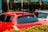 A parking lot or car wash area with two cars parked next to each other, one red and the other a lighter color. The red car has tinted windows, a roof rack, and a sticker on the back window. There are colorful signs and poles in the background with bushes in the foreground.
