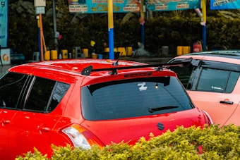 A parking lot or car wash area with two cars parked next to each other, one red and the other a lighter color. The red car has tinted windows, a roof rack, and a sticker on the back window. There are colorful signs and poles in the background with bushes in the foreground.