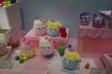 A collection of cute, colorful plush toys is displayed on a set of pink platforms. The toys have large, expressive eyes and resemble popular cartoon characters. Around the toys, there are small, colorful blocks, adding to the vibrant aesthetic.