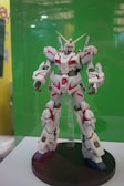 A robot toy model displayed on a black circular stand with intricate details, featuring a combination of white and red panels and gold accents. It stands against a plain green background, suggesting it's part of an exhibit or showcase.