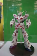A robot toy model displayed on a black circular stand with intricate details, featuring a combination of white and red panels and gold accents. It stands against a plain green background, suggesting it's part of an exhibit or showcase.