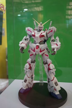 A robot toy model displayed on a black circular stand with intricate details, featuring a combination of white and red panels and gold accents. It stands against a plain green background, suggesting it's part of an exhibit or showcase.
