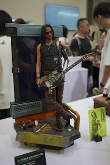 A detailed and intricately designed figurine of a male character leaning against a backdrop. The figure is wearing sunglasses, a tactical vest, and leather pants, holding a guitar. The base of the figure includes various elements like bottles, a pair of sunglasses, and a sign reading 'rock & place.' Several people are visible in the background, suggesting a convention or expo setting.