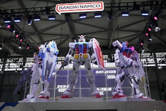 Three large, detailed mecha models stand prominently on a platform in a well-lit exhibition space. They are intricately designed, with various colors and accessories, each showcasing futuristic armor and weaponry. Overhead, a sign reads 'Bandai Namco,' and the background features structural metal beams with lights.