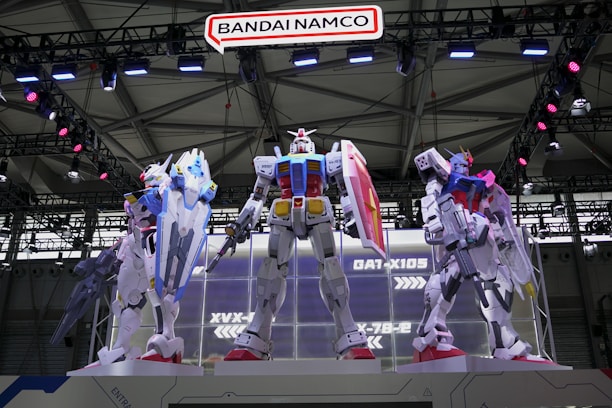 Three large, detailed mecha models stand prominently on a platform in a well-lit exhibition space. They are intricately designed, with various colors and accessories, each showcasing futuristic armor and weaponry. Overhead, a sign reads 'Bandai Namco,' and the background features structural metal beams with lights.