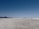 A vast, open salt flat stretches toward the horizon with distant mountain ranges visible under a clear blue sky. The expansive plain appears deserted except for a single vehicle and a few people near it, adding a sense of scale to the wide landscape.