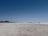 A vast, open salt flat stretches toward the horizon with distant mountain ranges visible under a clear blue sky. The expansive plain appears deserted except for a single vehicle and a few people near it, adding a sense of scale to the wide landscape.