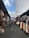 A vibrant, bustling market aisle is lined with stalls featuring a variety of colorful clothing, woven baskets, and textiles. Shoppers walk through the narrow path, shaded by large white tarps overhead. The ambiance suggests a lively, open-air environment.